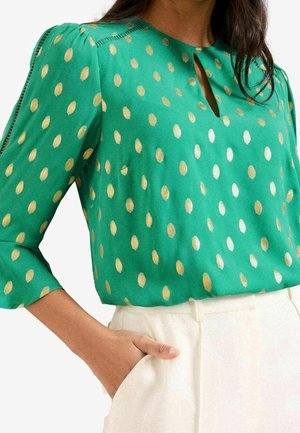 Kobieta ubrana w zieloną bluzkę w złote groszki z rękawami do łokcia, do tego wysokie białe spodnie, z ręką w kieszeni.