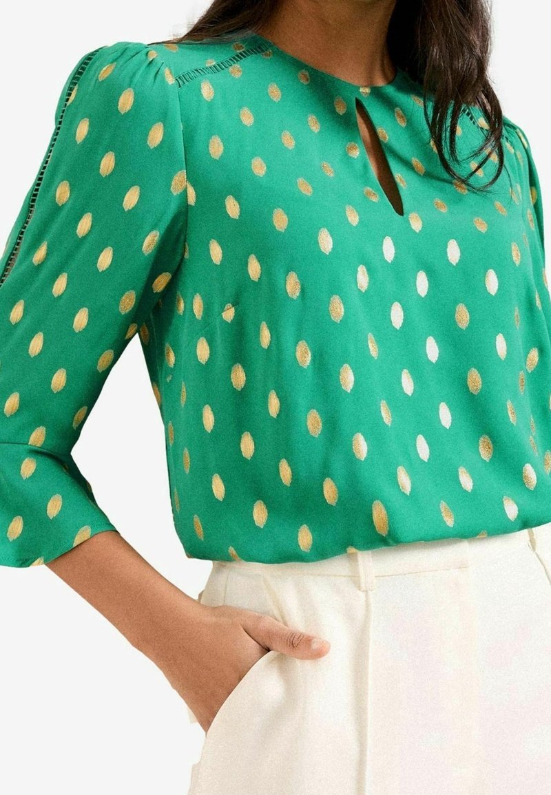Femme portant un chemisier vert à pois dorés avec des manches trois-quarts, associé à un pantalon blanc taille haute, main dans la poche.
