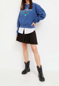 Sudadera azul con el texto "Bound Together" y diseño de cereza, superpuesta sobre una camisa blanca y combinada con una falda plisada negra y botas negras gruesas.