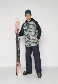 Σετ για σκι που περιλαμβάνει μια πράσινη καμουφλαρισμένη μπλούζα με κουκούλα, σκούρο παντελόνι και μαύρα μπότες σκι, κρατώντας ένα σκι με δεσίματα. Το σκουφάκι προσφέρει ζεστασιά.