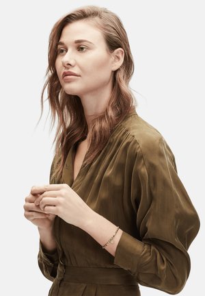 Olijfgroene satijnen blouse met geplooid ontwerp, lange mouwen en riem. Goudkleurige armband met ronde hanger om de pols. Glad textuur.