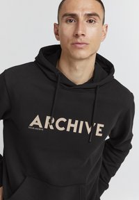 Sudadera negra con un bolsillo tipo canguro, capucha con cordón y gran texto en crema que dice "ARCHIVE". Hecha de un material suave con una textura lisa.
