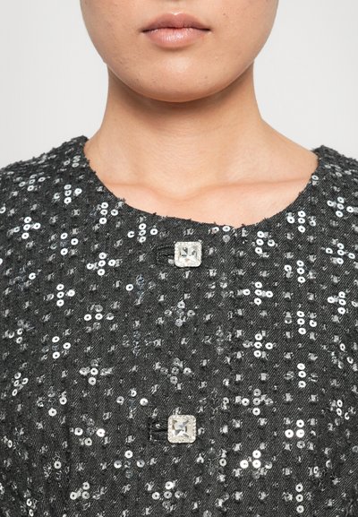 Close-up of a person's lower face and neck wearing a black sequined top with two square crystal buttons.