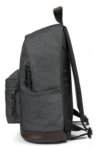 Mochila de tela gris con superficie texturizada, acentos negros, bolsillo lateral y base marrón. Cuenta con correas de hombro ajustables y un asa superior.
