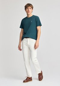 T-shirt a maniche corte color teal in un tessuto morbido, abbinato a pantaloni bianchi a gamba dritta e scarpe stringate marroni, su uno sfondo neutro.