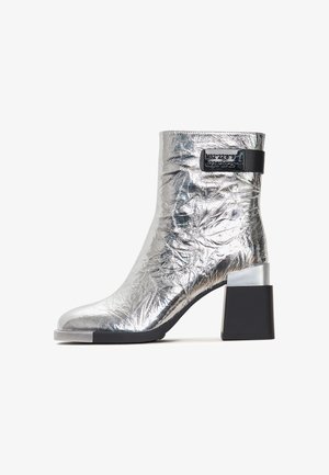Silberner Knöchelstiefel mit einer zerknitterten metallischen Oberfläche, ausgestattet mit einem kontrastierenden schwarzen Riemen und einem klobigen Blockabsatz mit einem silbernen Akzent.