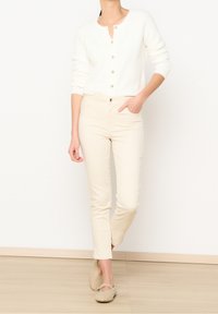 Vit stickad cardigan med guldknappar, tillsammans med beigefärgade figurnära byxor och ljusbruna slip-on skor med nitar.