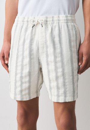 Mann trägt weiße Shorts mit schwarzen vertikalen Streifen und einem elastischen Bund mit einer gebundenen Kordel, steht mit entspannten Armen.