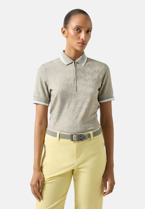 Femme portant un polo à manches courtes gris clair avec un motif de feuilles et un pantalon jaune pâle avec une ceinture tissée grise, posant devant un fond blanc.