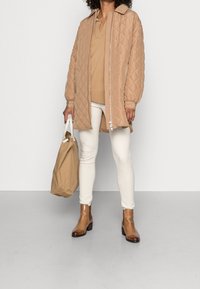 Veste matelassée marron clair avec fermeture à glissière sur le devant, manches bouffantes et col, associée à un pull beige, un pantalon blanc et des bottines marron.