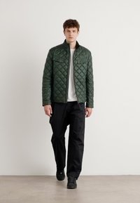 Veste matelassée verte avec un col haut, zip frontal et deux poches poitrine, associée à un pantalon et des chaussures noirs, se tenant sur un fond neutre.
