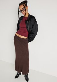 Kobieta w warkoczach, ubrana w czarną kurtkę bomber, bordowy top, brązową maksiskirtę, czarne botki na obcasie oraz okulary przeciwsłoneczne, pozująca na tle białej ściany.