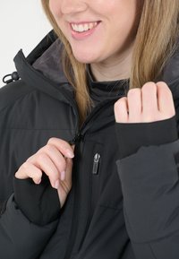 Svart isolerad jacka med hög krage och blixtlåsförslutning. Har hål för tumme, justerbar huva och ficka med dragkedja. Mjuk textur synlig.