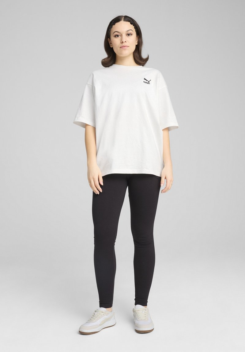 Biała, oversize'owa koszulka bawełniana z małym czarnym logo, zestawiona z czarnymi legginsami i białymi sneakersami. Prosty design, luźny krój.