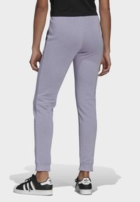 Osoba oblečená v světle fialových přiléhavých teplácích a černých teniskách Adidas s bílými pruhy, stojící na jednolitém pozadí.