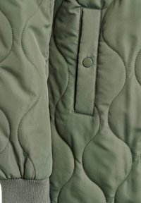 Zaļas šūtas jaka audums ar izliektiem šuvju rakstiem, kam ir atloks ar kabatu un ribošanas aproce piedurknes galā.