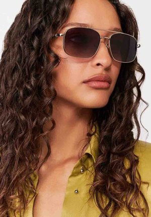 Kobieta z długimi kręconymi włosami, nosząca duże kwadratowe ciemne okulary przeciwsłoneczne i oliwkowo-zieloną koszulę z kołnierzykiem, zwrócona w prawo.