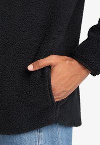 Fekete fleece dzseki textúrált felülettel, oldalsó zsebbel és húzott ujjal. Az anyaga puha és kényelmes.