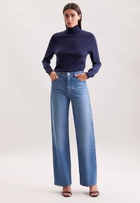 Marineblauer Rollkragenpullover kombiniert mit hoch taillierter, weit geschnittener Jeans in Hellblau. Glatte Stoffstruktur und klassische Knopfverschluss.