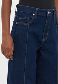Mörkblå högmidjade denimjeans med kontrasterande sömmar, bärs med en svart topp, visar en närbild av midjan och vänster hand.