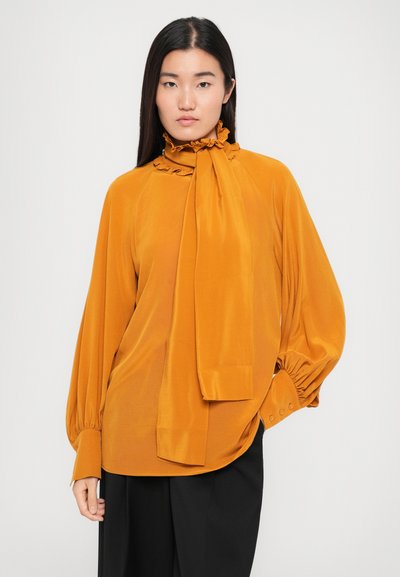 Blusa gialla senape realizzata in tessuto morbido, con colletto arricciato e dettaglio con fiocco, maniche lunghe e arricciate, dalla vestibilità morbida.