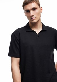 Czarny polo z teksturowanym wykończeniem, krótkimi rękawami i dekoltem w serek. Prosty wzór bez widocznych logo czy wzorów.