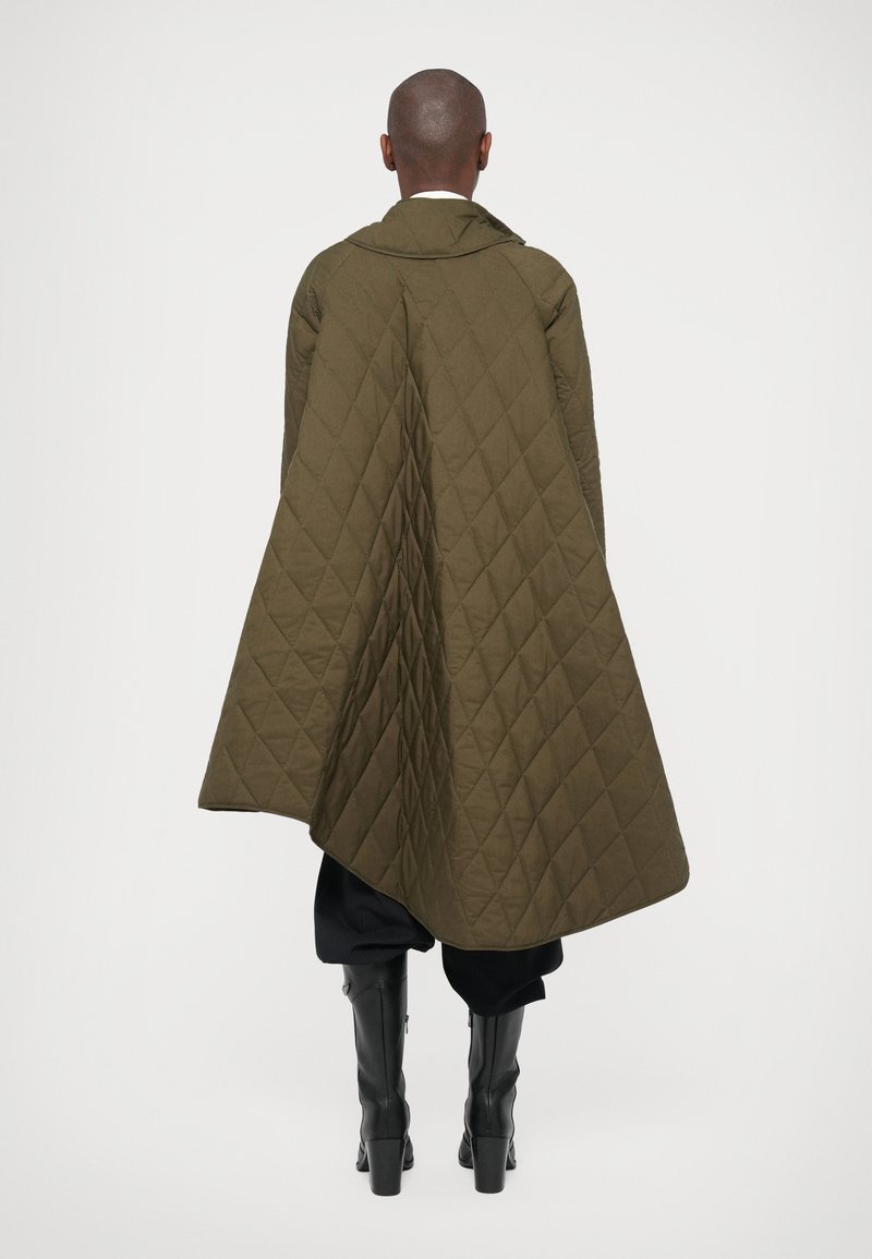 Personne portant une longue cape matelassée de couleur olive avec des bottes noires, se tenant dos à un fond blanc uni.