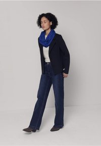 Marineblauw jasje, wit topje, blauwe sjaal en donkere denim flared jeans. Het figuur draagt bruine enkellaarsjes en toont een casual, gelaagde look.