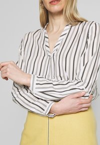 Femme portant un chemisier blanc avec des rayures verticales noires et une jupe jaune avec une fermeture éclair devant, bras croisés à la taille.