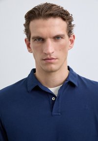 Hombre con cabello rizado castaño y ojos azules que lleva una camisa de color azul marino con cuello sobre una camiseta blanca, contra un fondo suave y claro.