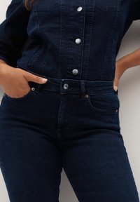 Persona con chaqueta de mezclilla azul oscuro y jeans con las manos en los bolsillos, mostrando los detalles frontales del botón y la cremallera.