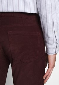 Gros plan sur une personne portant un pantalon en velours côtelé bordeaux et une chemise blanche à rayures avec boutons, montrant la poche arrière et la main droite.