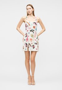 Jeune femme portant une robe blanche sans manches avec des motifs abstraits colorés et animaliers, debout les mains dans le dos, fond neutre.