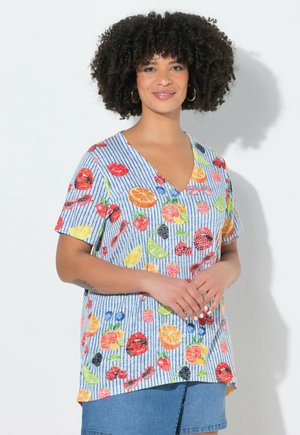 Kvinde med krøllet hår iført store øreringe og en stribet V-hals trøje med farverige frugtmønstre, stående med hænderne foldet.