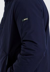 Veste bleu marine avec un tissu texturé, ornée d'un patch logo RNT23 bien visible sur la manche et d'une poche zippée sur le côté.