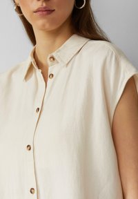 Beige Hemd mit kurzer Arm, weichem Stoff, klassischem Kragen und braunen Knöpfen entlang der Vorderseite.