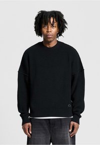 Black ribbed knit sweater with a round neckline, dropped shoulders, and long sleeves. Features a subtle logo on the hem and layered look.