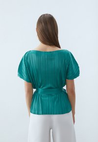 Blusa plissettata color turchese con scollatura ampia e maniche corte, cintrata in vita con una fascia. Texture liscia e caduta morbida.