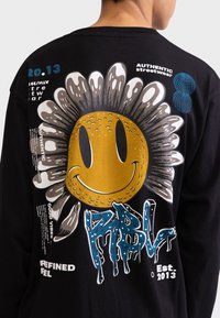 Schwarzes Langarmshirt mit einem großen lächelnden Sonnenmotiv in Gold, silbernen Blütenblättern, blauen Akzenten und weißen Textelementen.