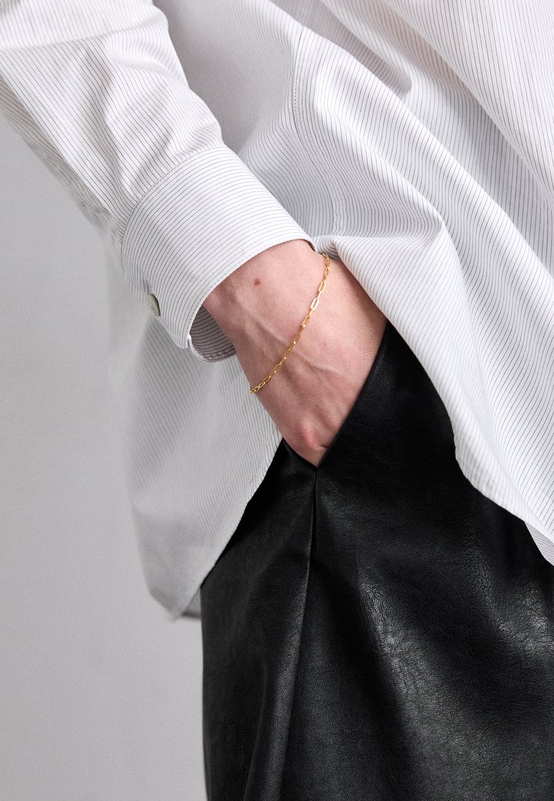 Bela črtasta srajca sovičastimi rokavi, v kombinaciji z gladko črno usnjeno krilo. Na zapestju je zlatih verižnih zapestnica.