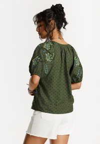 Blouse en coton vert avec des manches bouffantes, ornée de broderies florales en bleu et jaune. Dots texturés sur l'ensemble. Associée à un short blanc.