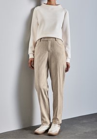 Ljus krämfärgad sweatshirt med hög krage, kombinerad med beiga, texturerade byxor som har en enda knapp och sidfickor. Vita sneakers med grå detaljer.