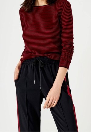 Femme portant un haut à manches longues rayé rouge et noir associé à un pantalon noir à cordon avec des bandes rouges latérales.