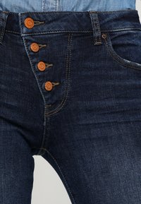 Mörkblå denimjeans med fem knappar i midjan, orange detaljer, sydda dekorationer och en slim-fit design.