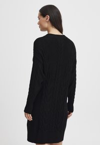 Abito nero lavorato a maglia con motivo a trecce testurizzato, maniche lunghe e polsini a coste. Lunghezza a metà coscia con una vestibilità rilassata e scollatura rotonda.