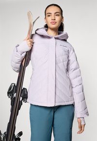 Lyselilla isoleret jakke med diamantmønster, udstyret med hætte og lynlåslommer. Model holder ski og skibindinger.