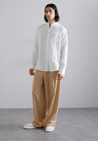 Camicia bianca in lino a maniche lunghe con taschino sul petto, abbinata a pantaloni beige a gamba larga e sneakers bianche su un pavimento grigio.