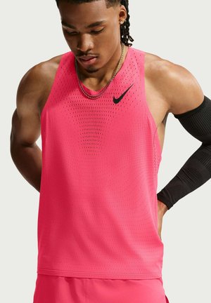 Atletische man met gevlochten haar, dragend een roze mouwloos Nike-topje en bijpassende korte broek, kijkt naar beneden met handen achter zijn rug.