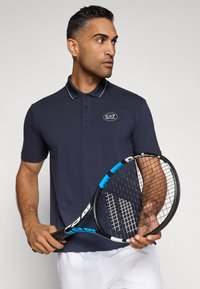 Námornícky polo tričko so slim fit strihom, bielymi pruhmi na golieri a logom EA7; model drží modrú a čiernu tenisovú raketu.
