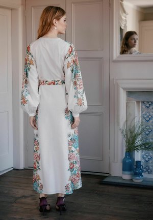 Woman in a white floral long dress with puffed sleeves stands in a room, facing a wall mirror reflecting her face.